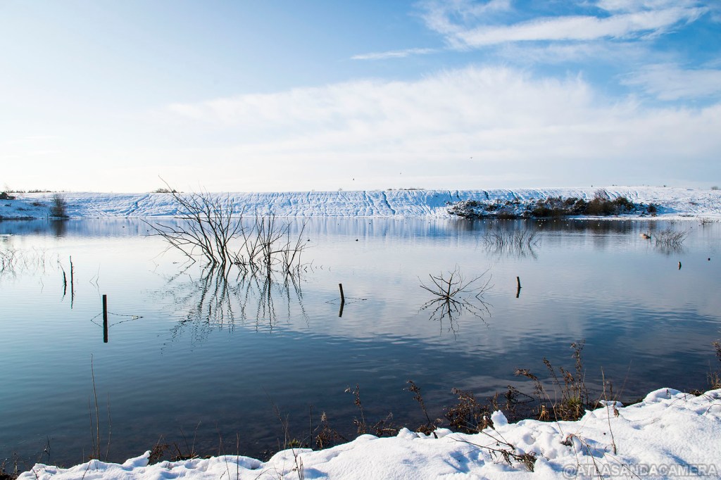 A blue sky over the bottom lagoon at Newstead and Annesley Country Park. The ground around it is covered in snow.