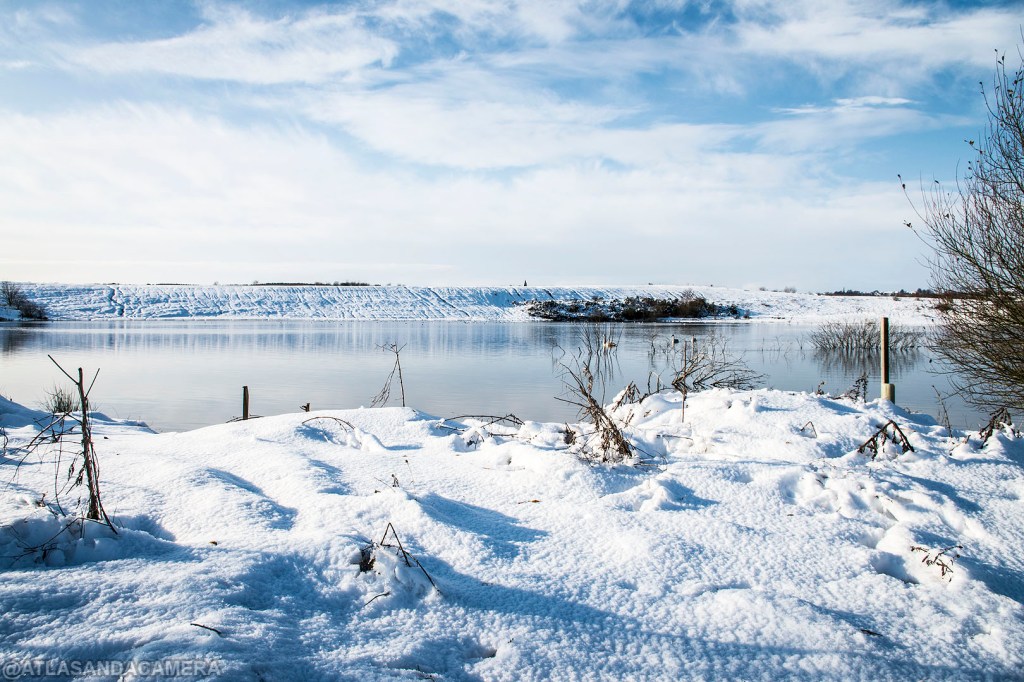 A blue sky over the bottom lagoon at Newstead and Annesley Country Park. The ground around it is covered in snow.
