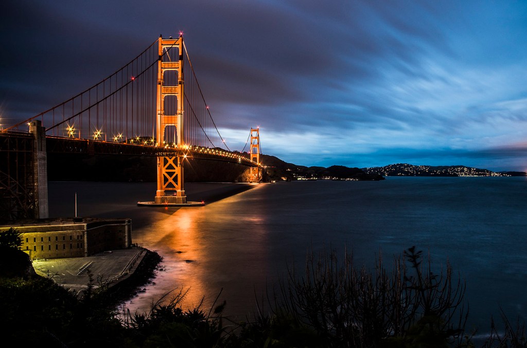 Golden Gate Bridge at night. The red bridge is reflected on the water below. There are city lights on hills in the distance.