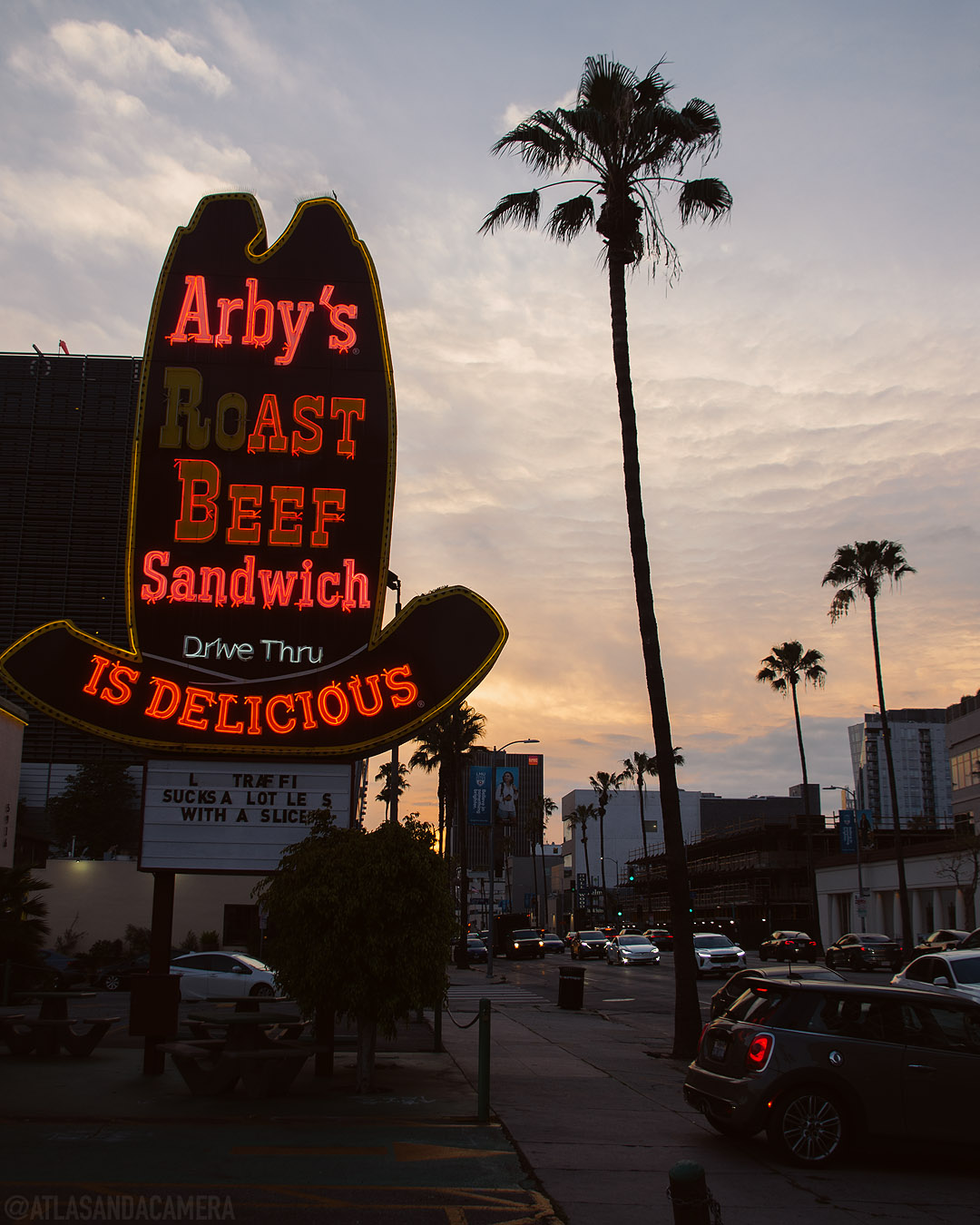 A neon red Arby's sign on the palm-lined Sunset Boulevard at sunset.