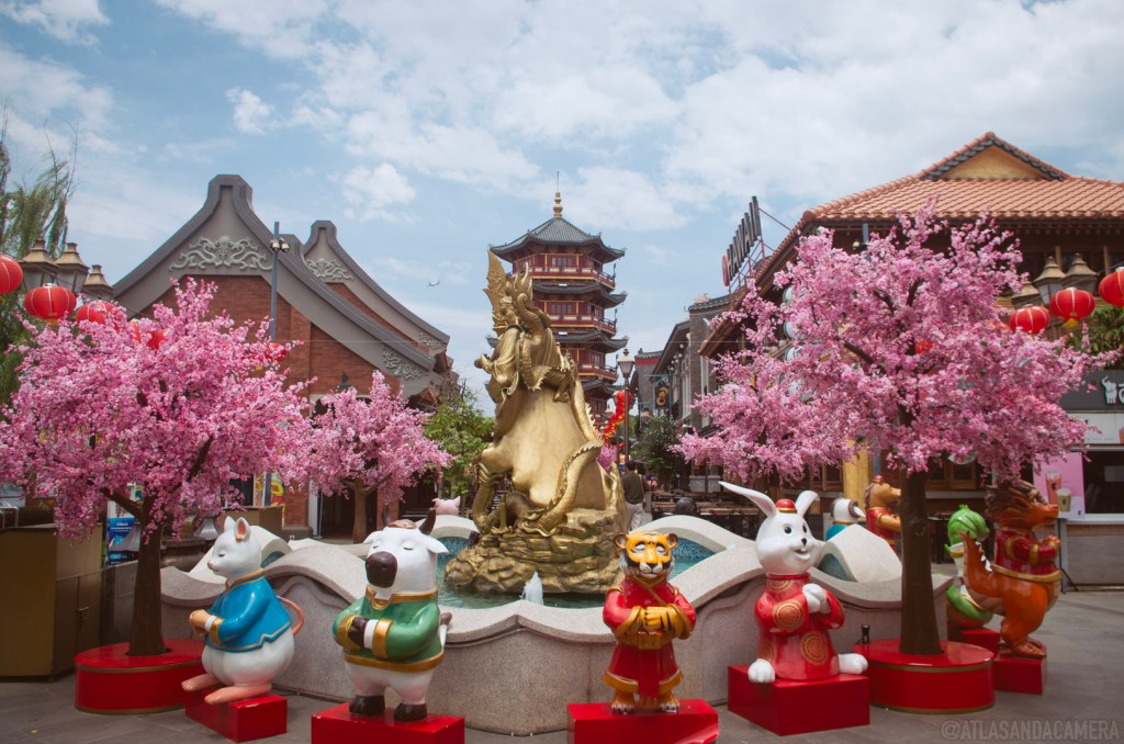 Pantjoran (Chinatown) PIK in Jakarta. Trees with pink blossom and zodiac sculptures surround a fountain in front of a pagoda.