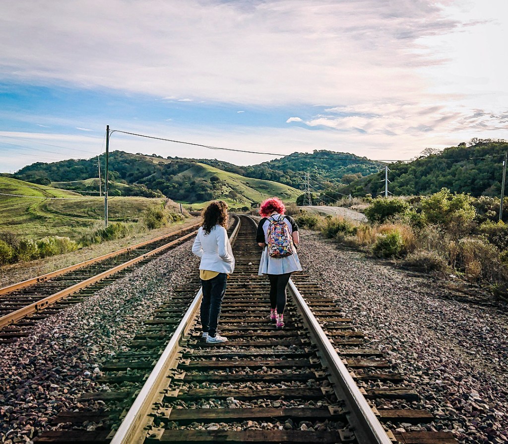 Two people on train tracks at Christie Road, surrounded by green hills. The sky is blue.