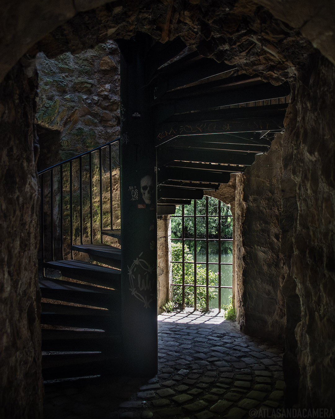 Inside one of the ruined casemates by the riverside in Luxembourg. In the dimly lit space, a metal spiral staircase has been added and a barred window looks out onto the river.