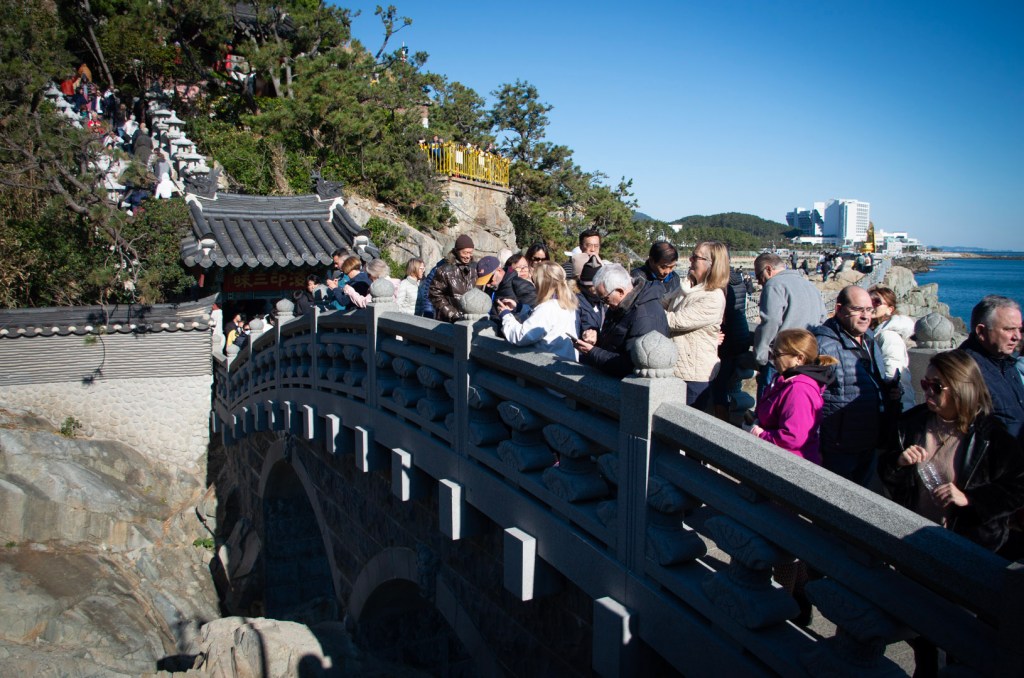 Crowds of tourists in winter coats on an arched bridge at Haedong Yonggungsa Temple, surrounded by cliffs and the ocean in the distance.