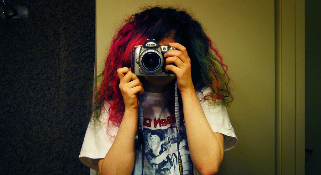 A reflection of a woman in a white t-shirt holding a silver Nikon camera in a dimly-lit hotel room.