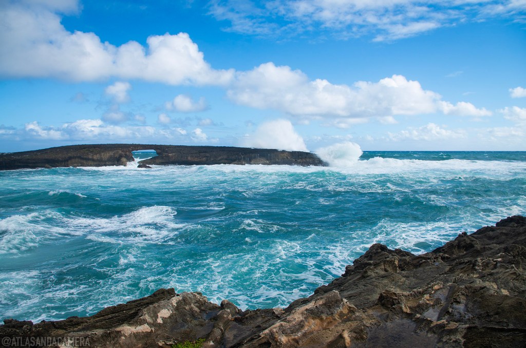 The view of Kukuihoolua Island from the rocky brown cliffs at Laie Point State Wayside. A wave is crashing over the island with its rock arch. The sky is blue with some clouds.
